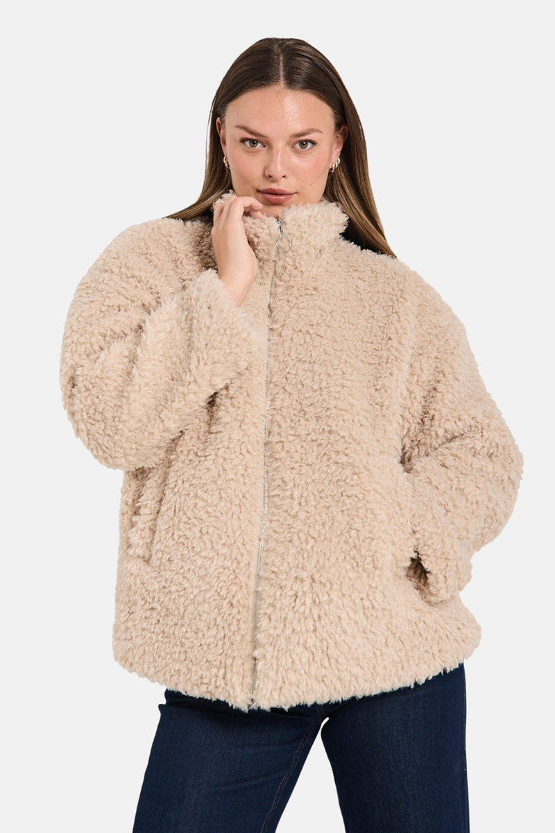 Veste courte beige en fausse fourrure de FREEQUENT, avec fermeture éclair et poches, portée avec un jean.
