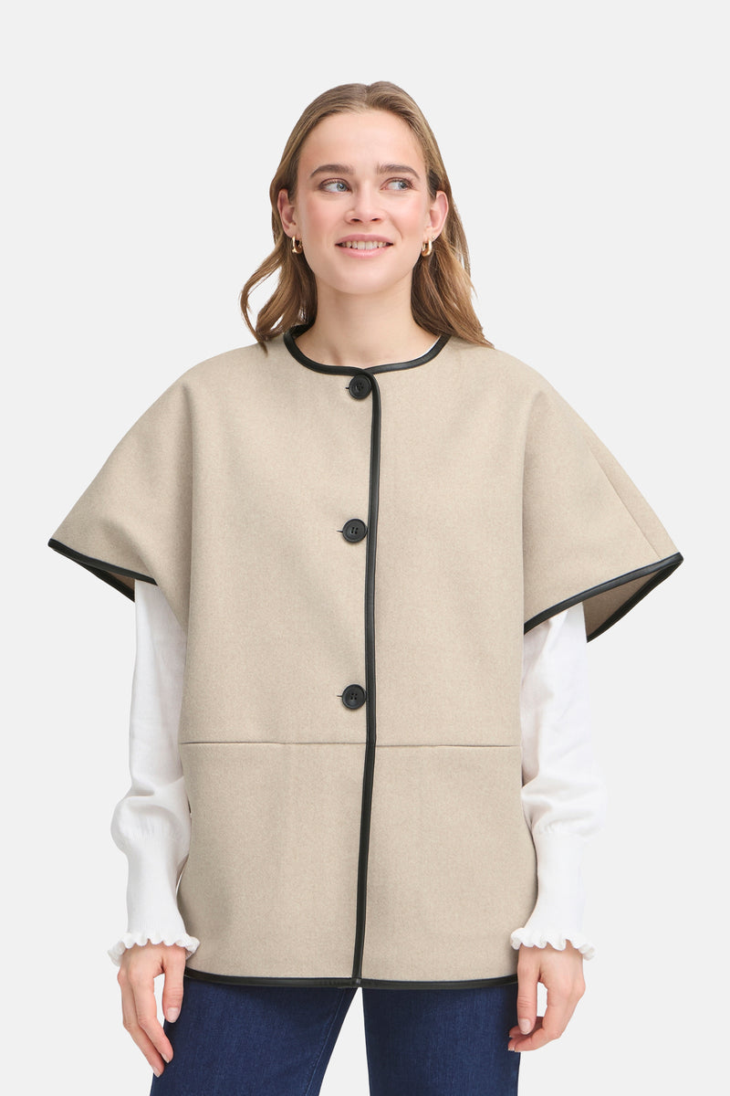 Veste beige, de Fransa, avec boutons et col en cuir noir, portée sur un chemisier blanc à manches longues.
