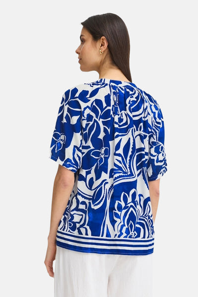 Blouse à manches courtes bleue de Fransa ornée d'un motif floral contrastant, portée avec un pantalon blanc.