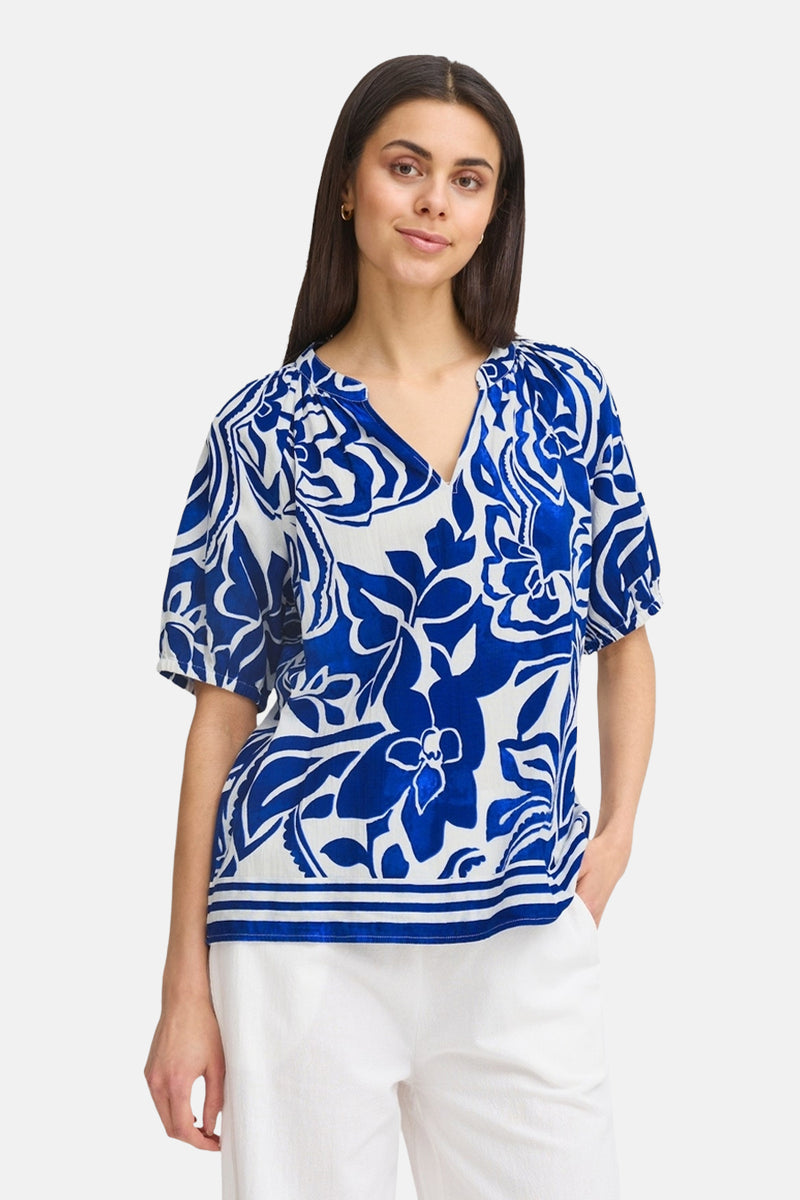 Chemisier bleu à fleurs by Fransa®, avec manches courtes et bordure rayée, porté avec un pantalon blanc.
