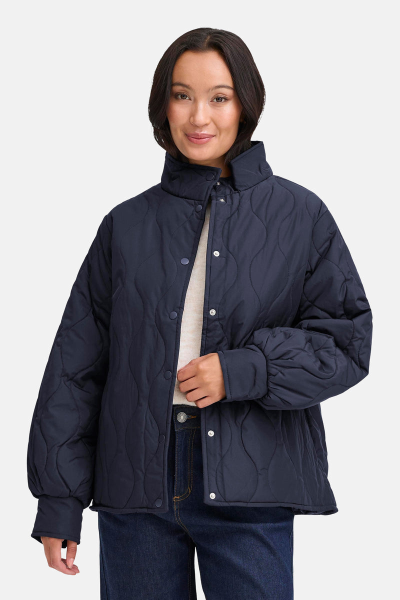 Veste courte bleue de Fransa, à motif matelassé et boutons-pression, portée avec un pull beige et un jean foncé.