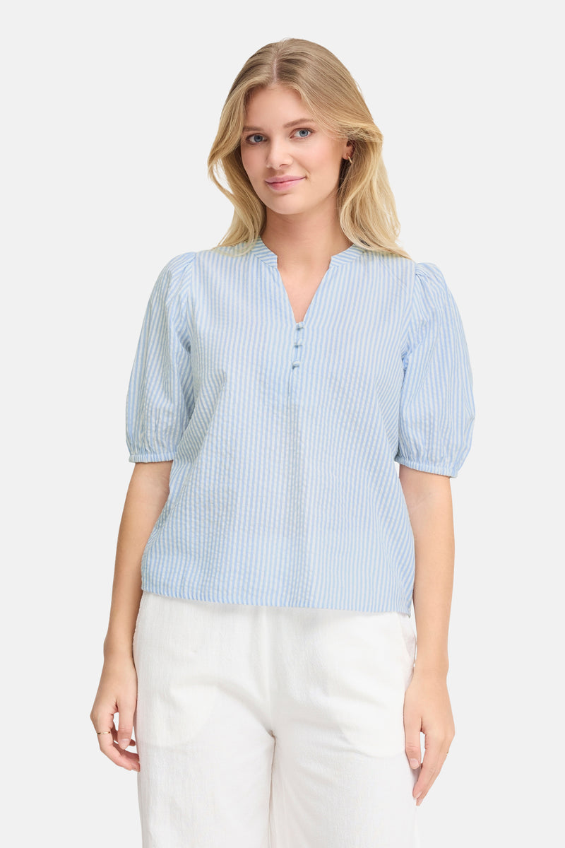 Blouse à manches courtes bleue rayée de Fransa, avec boutons et manches bouffantes, portée avec un pantalon blanc.