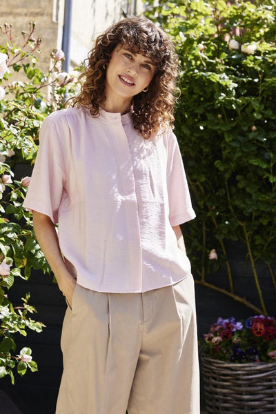 Chemise rose à manches courtes de Fransa, col mao, texture crêpée, portée avec un pantalon beige plissé.