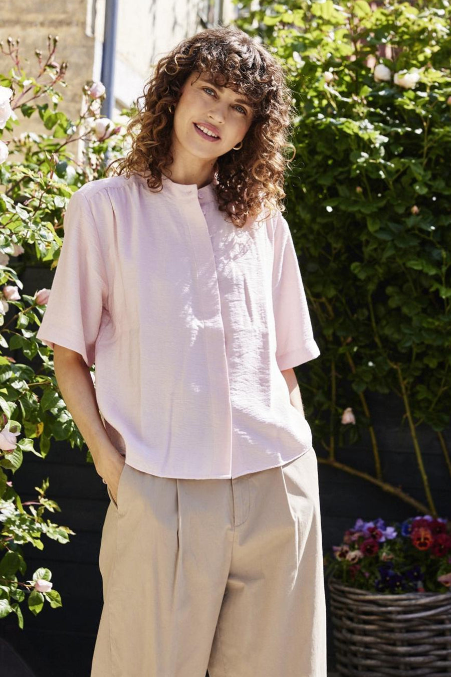 Chemise rose à manches courtes de Fransa, col mao, texture crêpée, portée avec un pantalon beige plissé.