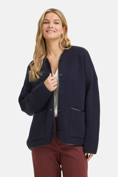 Gilet bleu marine en maille épaisse par Fransa®, boutonné avec poches et bordure contrastante, porté sur un haut blanc ajouré et pantalon bordeaux.