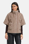 Beige fuzzy short coat with turtleneck and front pockets, worn over dark long-sleeve top and black shiny pants, front view