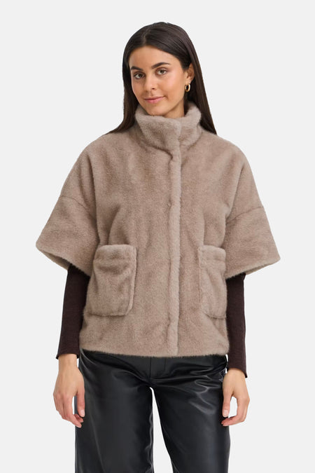 Beige fuzzy short coat with turtleneck and front pockets, worn over dark long-sleeve top and black shiny pants, front view