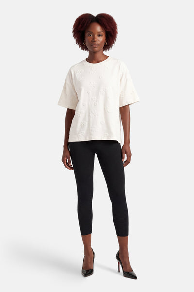 T-shirt blanc à manches courtes de FREEQUENT, motif floral en relief, porté avec un legging noir et des escarpins vernis.