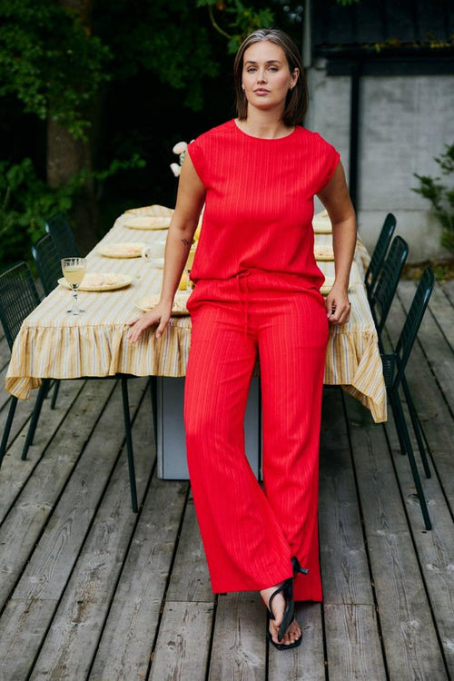 Top sans manches rouge de FREEQUENT, porté avec un pantalon coordonné, devant une table dressée sur une terrasse en bois.
