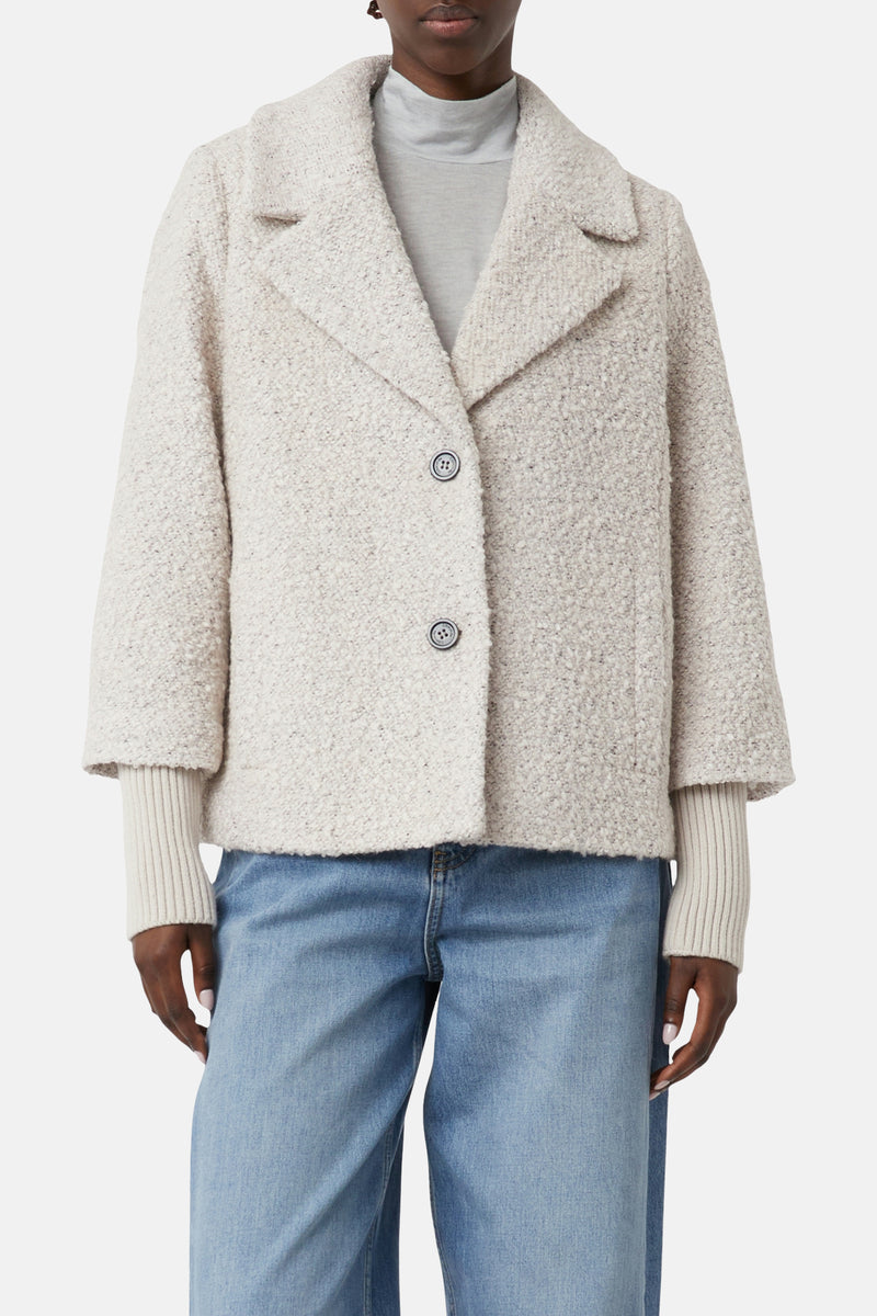 Korte beige jas van COMMA met bouclétextuur, gedragen over een coltrui en een blauwe jeans.