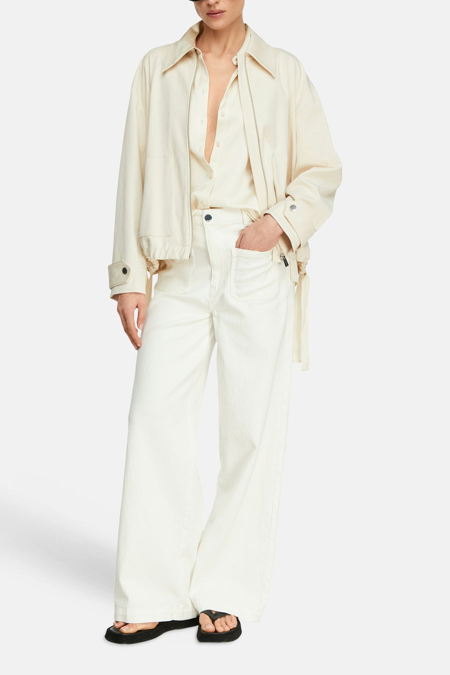 Korte beige jas van COMMA, met ritssluiting en grote zakken, gedragen over een wit overhemd en een witte broek.