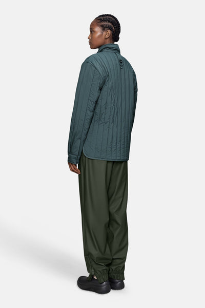 Dark green quilted women's puffer jacket with vertical stitching, side view, worn with olive pants and black shoes