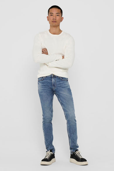 Jean slim bleu de ONLY & SONS, porté avec un pull blanc à manches longues et des baskets noires.