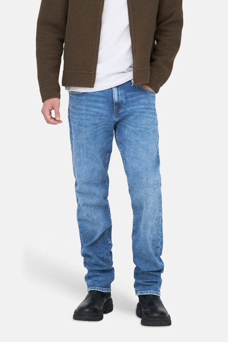 Jeans droit bleu moyen de ONLY & SONS, avec des jambes droites et amples, porté avec un t-shirt blanc et une veste marron.
