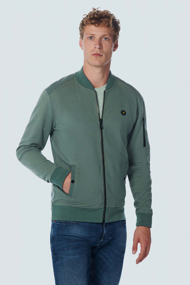 Green textured zip-front sweat gilet with ribbed cuffs and waistband, featuring side pockets and a small hexagonal logo on the chest, worn over a light green tee with blue jeans.