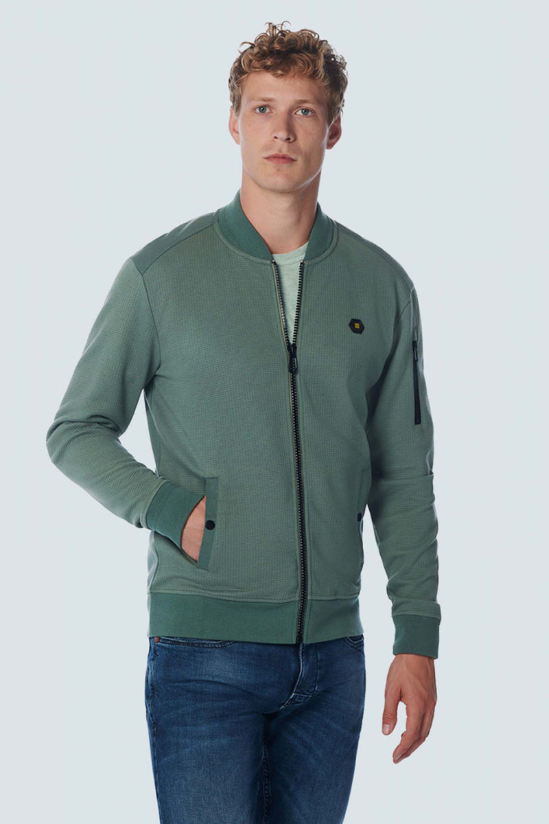 Green textured zip-front sweat gilet with ribbed cuffs and waistband, featuring side pockets and a small hexagonal logo on the chest, worn over a light green tee with blue jeans.