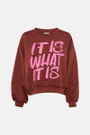Rust-colored crewneck sweatshirt with bold pink "IT IS WHAT IT IS" graphic, relaxed fit, ribbed cuffs and hem, front view
