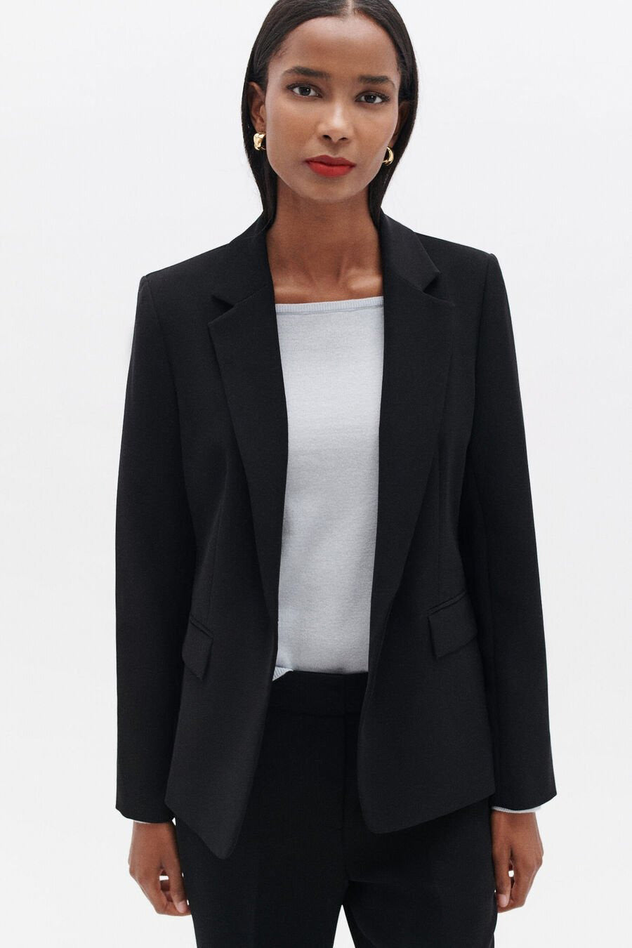 Blazer noir par CAROLL porté sur un top gris, avec des boucles d'oreilles dorées.