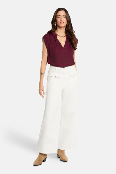 Chemisier bordeaux sans manches de Morgan de Toi, avec col en V et porté avec un pantalon blanc et des bottines marron.
