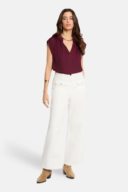 Chemisier bordeaux sans manches de Morgan de Toi, avec col en V et porté avec un pantalon blanc et des bottines marron.
