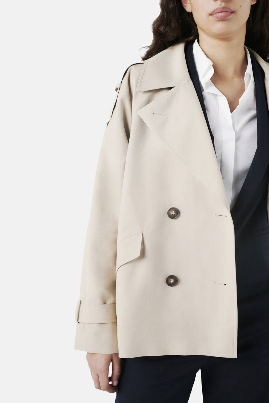 Veste courte beige de Caroll avec double boutonnage et manches à revers, portée sur une chemise blanche et un tailleur marine.
