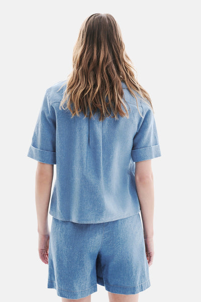 Chemisier denim bleu clair à manches courtes de CAROLL, vue arrière, avec cheveux longs et ondulés.
