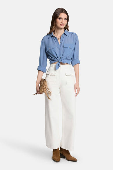 Blauwe blouse (lange mouwen) van Morgan de Toi, geknoopt in de taille, gedragen met een witte broek en bruine enkellaarsjes.