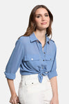 Blauwe blouse met lange mouwen van Morgan de Toi, met zakken, geknoopt aan de voorkant, gedragen met een witte broek.