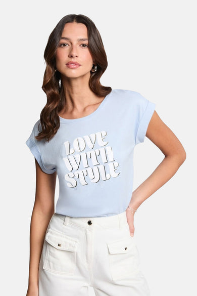T-shirt bleu à manches courtes de Morgan de Toi, avec lettrage blanc, porté avec un pantalon blanc.
