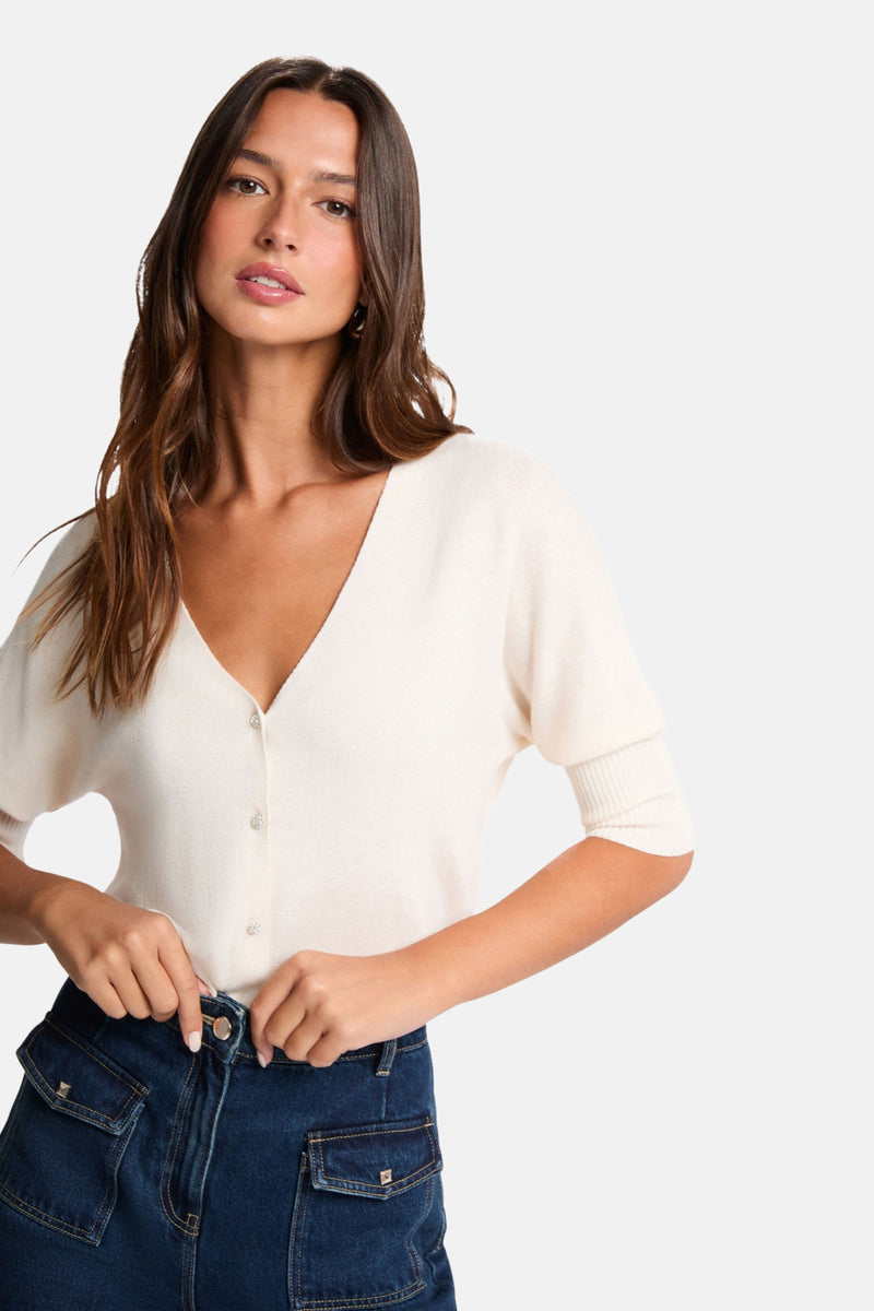 Cardigan écru de Morgan de Toi avec manches courtes et boutons décoratifs, porté avec un jean taille haute.
