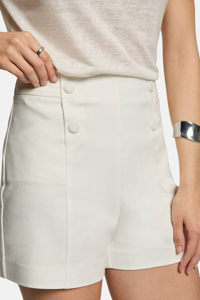 White high-waisted shorts with front buttons and side seam detailing, worn with a neutral top and silver cuff bracelet, front view.