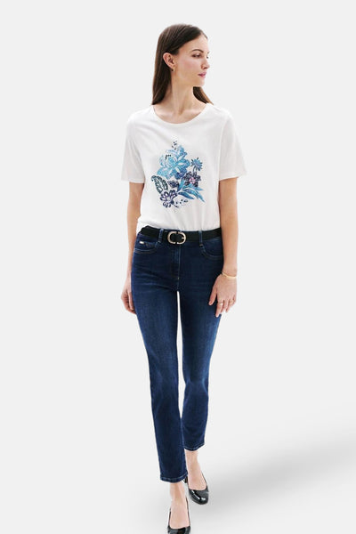 T-shirt blanc à manches courtes de CAROLL, avec un motif floral bleu sur le devant, porté avec un jean et une ceinture noire.
