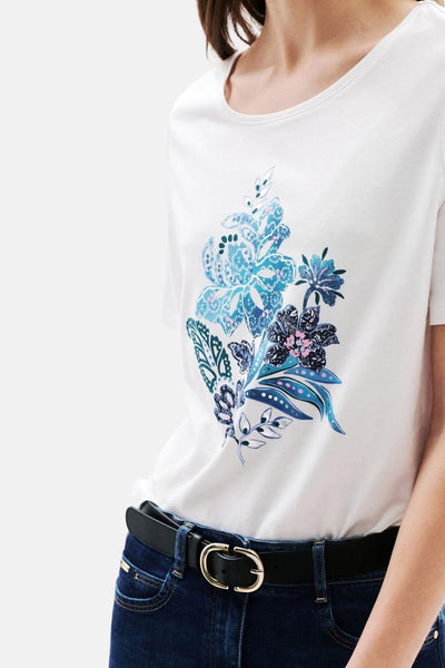 T-shirt blanc à manches courtes de CAROLL, avec motif floral bleu sur le devant, porté avec un jean et une ceinture noire.
