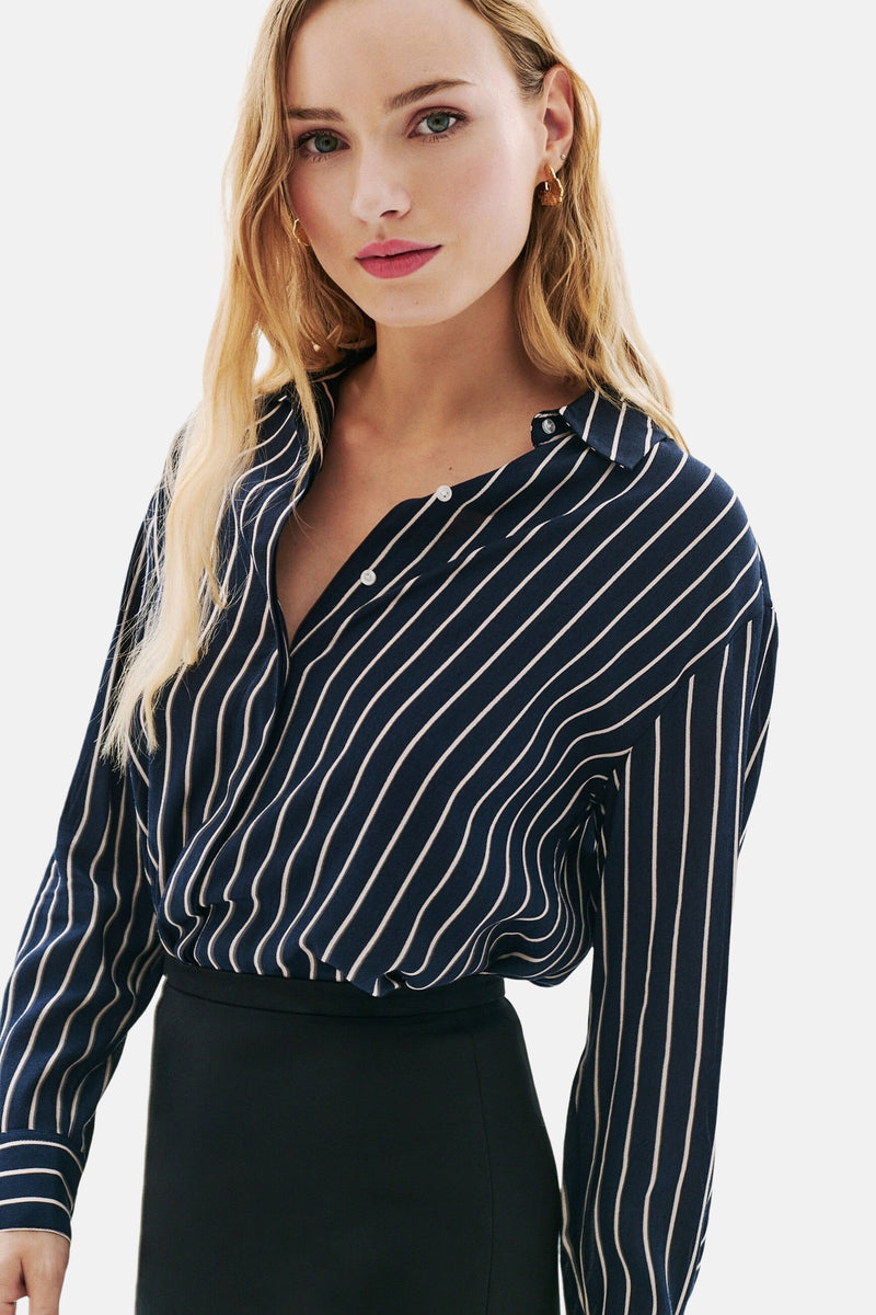 Blauwe gestreepte blouse met lange mouwen van CAROLL, gedragen met een zwarte rok, gedeeltelijk opengeknoopt en versierd met gouden oorbellen.
