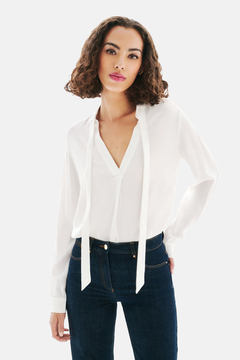 Witte blouse met lange mouwen van CAROLL, met V-hals en hangende strikjes, gedragen met een donkerblauwe jeans.