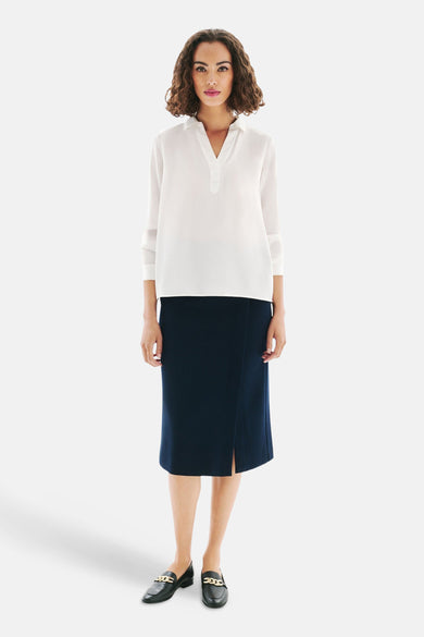 Blouse blanche à manches longues de CAROLL, portée avec une jupe bleu marine et des mocassins noirs à chaîne dorée.