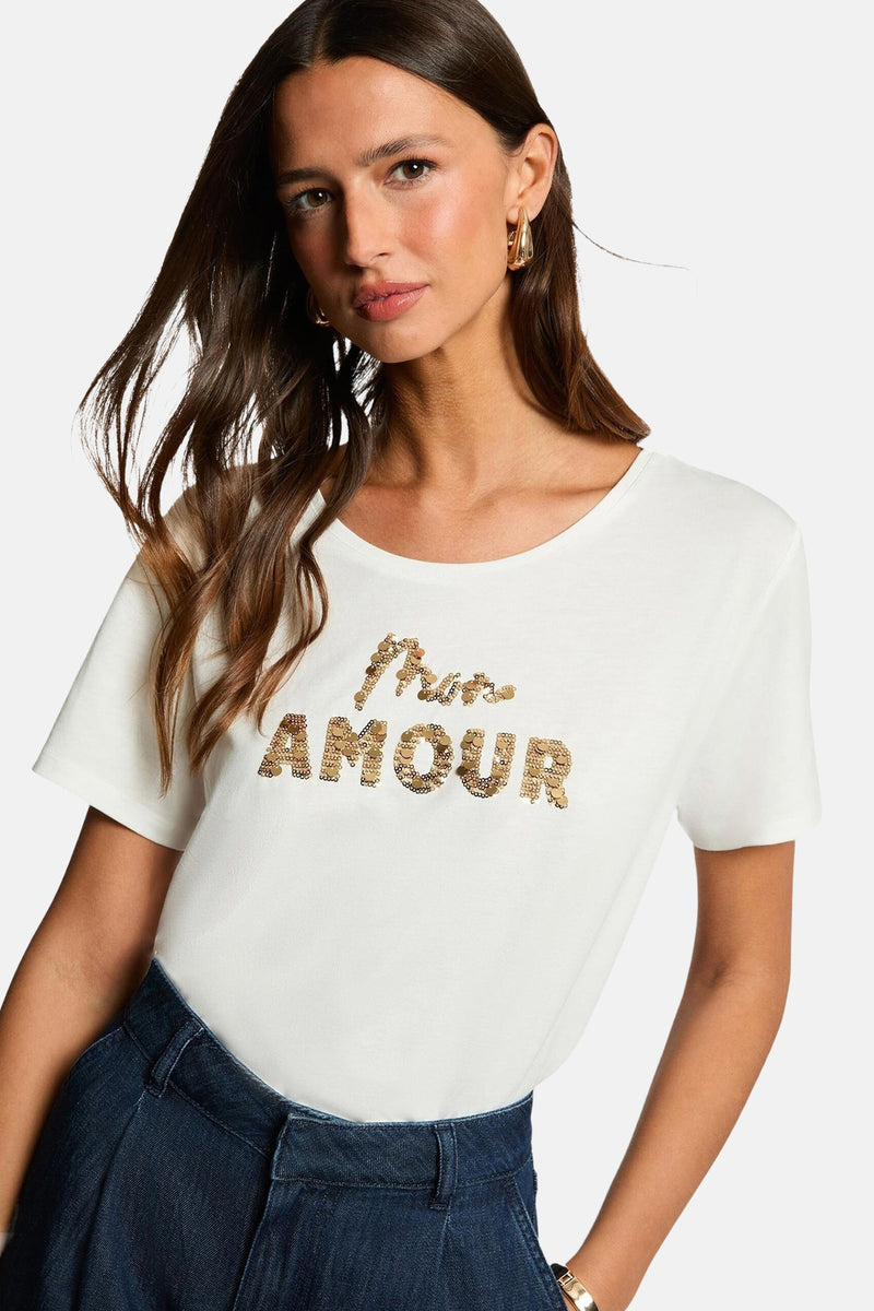 T-shirt à manches courtes blanc de Morgan de Toi avec inscription dorée à sequins, porté avec un pantalon.
