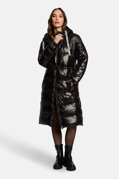 Veste longue noire de Morgan, brillante, avec capuche et portée avec des bottines noires et des collants noirs.
