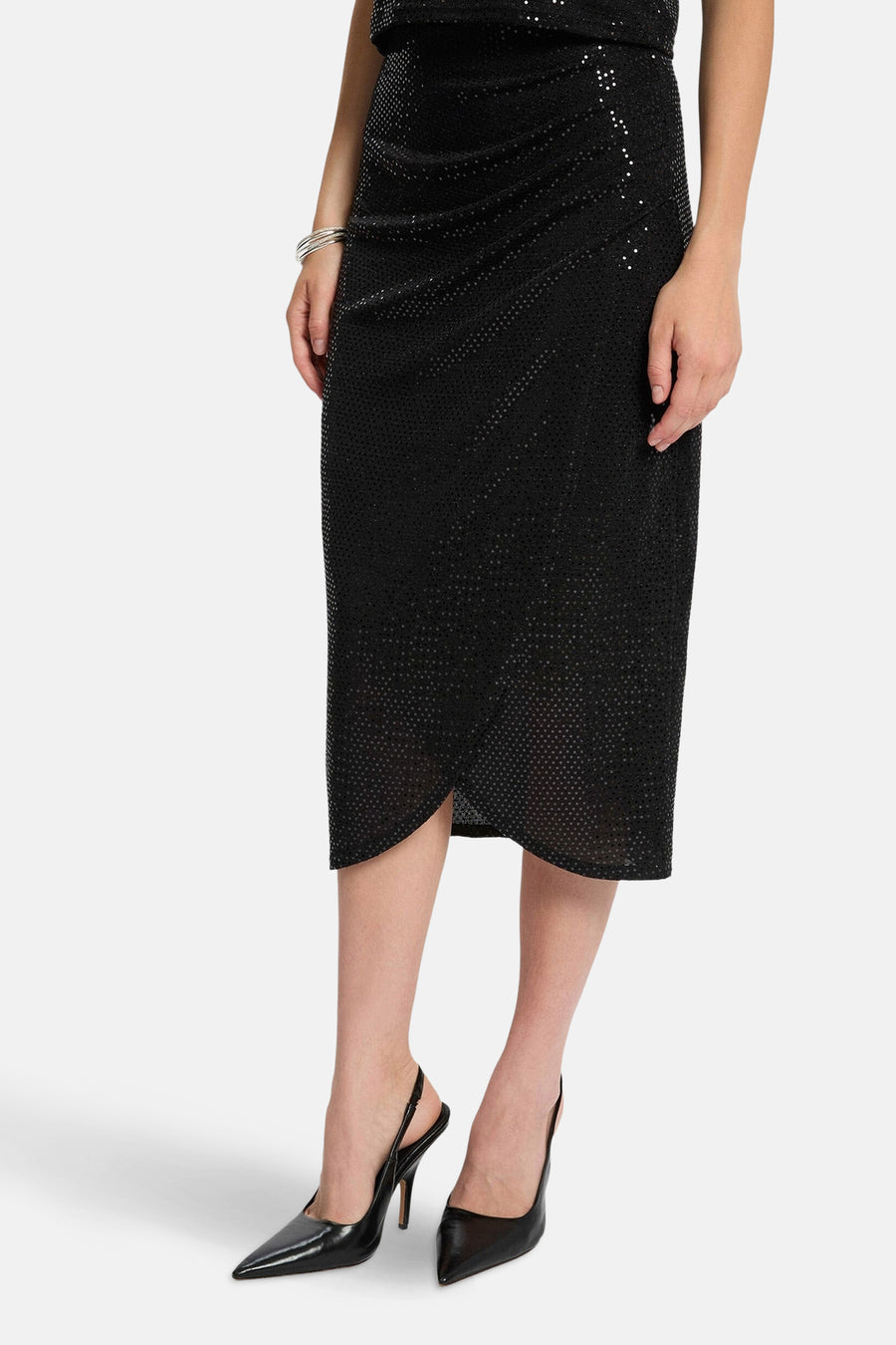 Jupe noire mi-longue de Morgan de Toi, coupe portefeuille drapée et détail pailleté, portée avec des escarpins noirs à talons aiguilles.