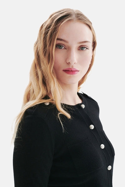 Cardigan noir de CAROLL avec des boutons argentés, porté avec des cheveux blond ondulés.