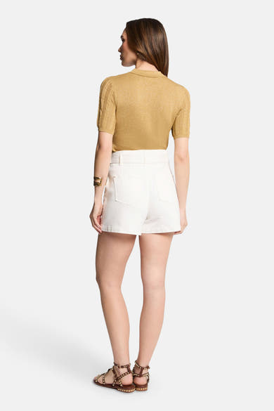Pull à col rond jaune de Morgan de Toi, manches courtes et texture tricotée, porté avec un short blanc et des sandales cloutées.