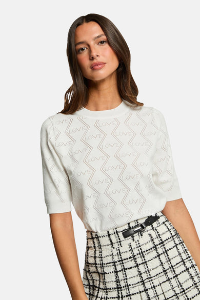 Pull à col rond blanc de Morgan de Toi, avec un motif ajouré « LOVE » et une jupe à carreaux.
