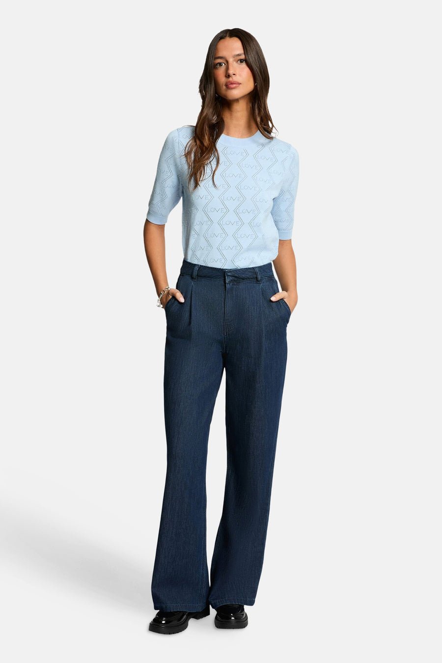 Ronde hals trui van Morgan de Toi in het blauw met ruitvormig 'LOVE' patroon, korte mouwen, gedragen over een wijde jeans en lakleren schoenen.