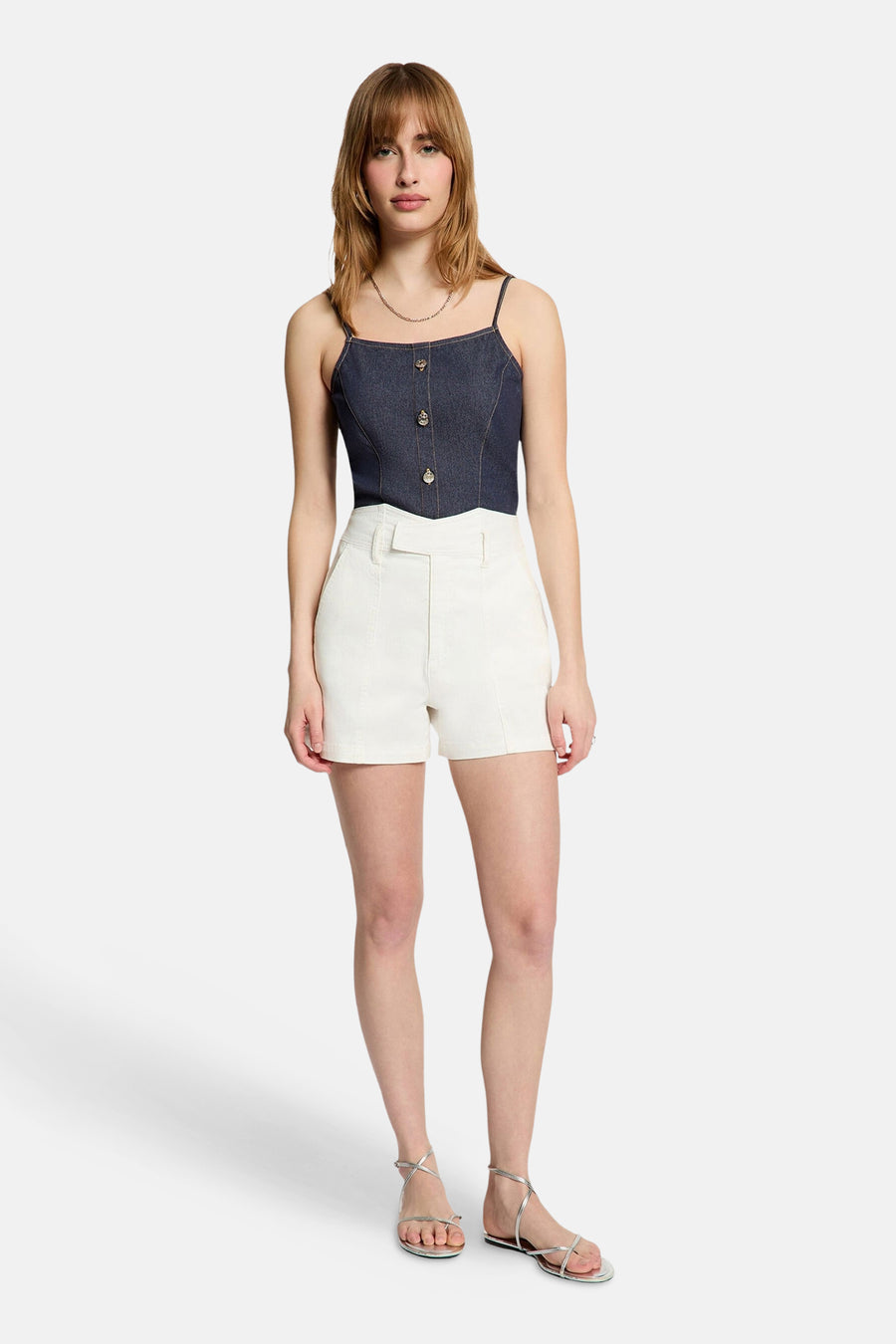 Top en jean bleu foncé de Morgan de Toi avec bretelles fines, boutons fantaisie et surpiqûres claires, porté avec un short écru et des sandales argentées.