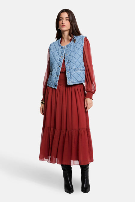 Veste sans manches en denim bleu clair de Morgan de Toi, matelassée en losanges, portée sur une robe longue brique et des bottes noires.
