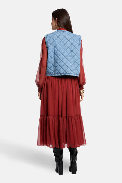 Veste sans manches en denim bleu clair de Morgan de Toi, matelassée avec motif losanges, portée sur une robe longue rouge et des bottes noires.
