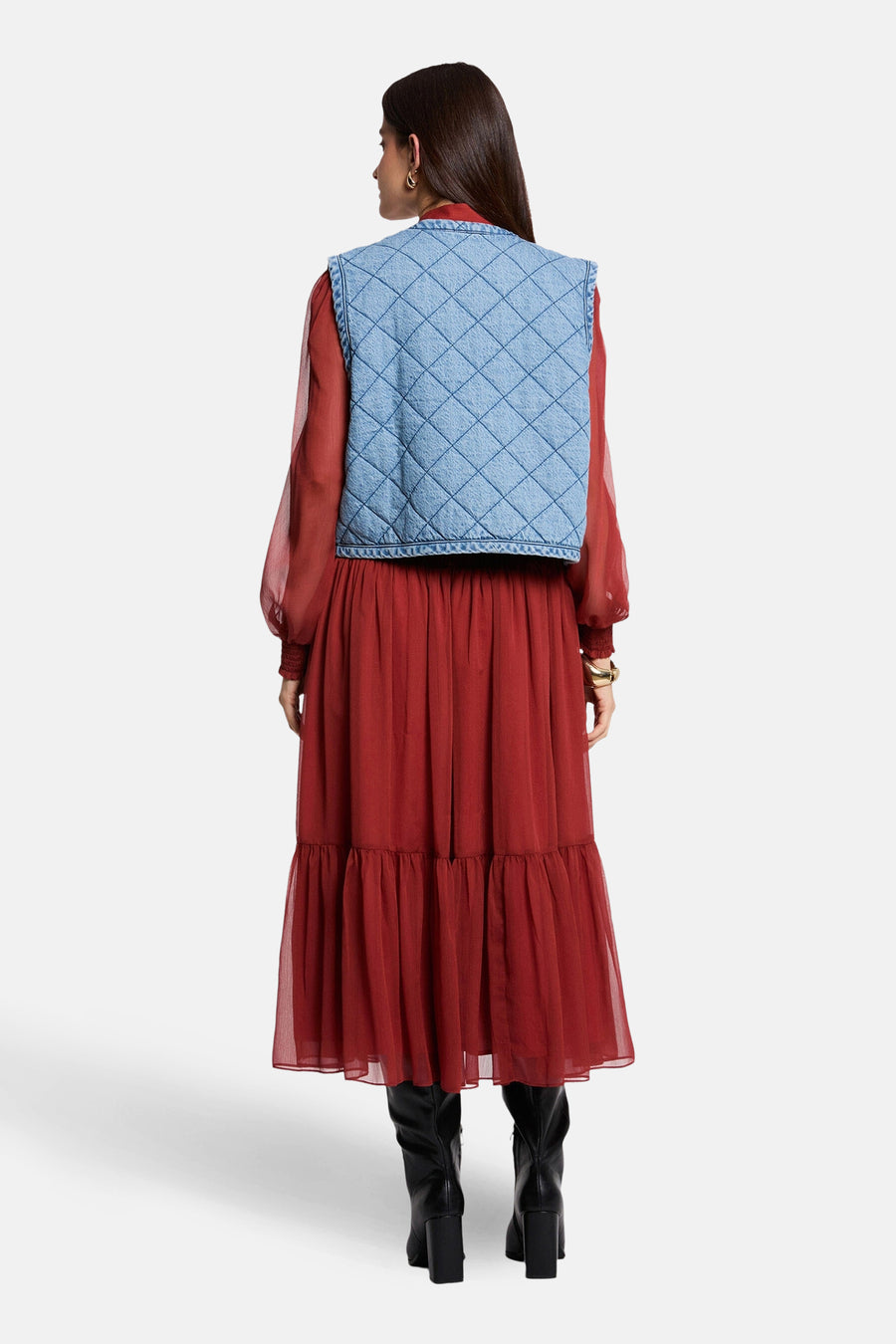 Veste sans manches en denim bleu clair de Morgan de Toi, matelassée avec motif losanges, portée sur une robe longue rouge et des bottes noires.
