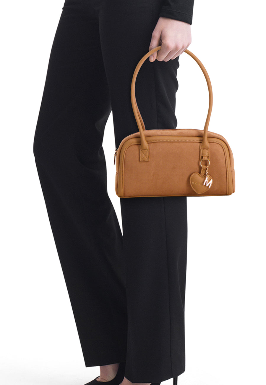 Cognac suede handbag with rounded top handles and heart-shaped charm, worn by model in black pants, side view.