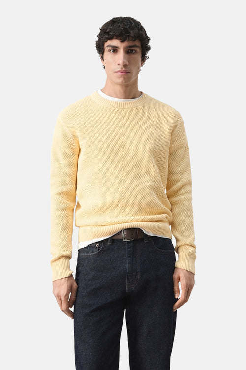 Pull à col rond jaune de MANGO, avec une texture tricotée et porté avec un jean foncé et une ceinture marron.