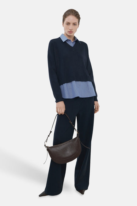 Pull col V bleu par MANGO, porté sur une chemise rayée bleu clair, avec un pantalon bleu et un sac à main en cuir marron.